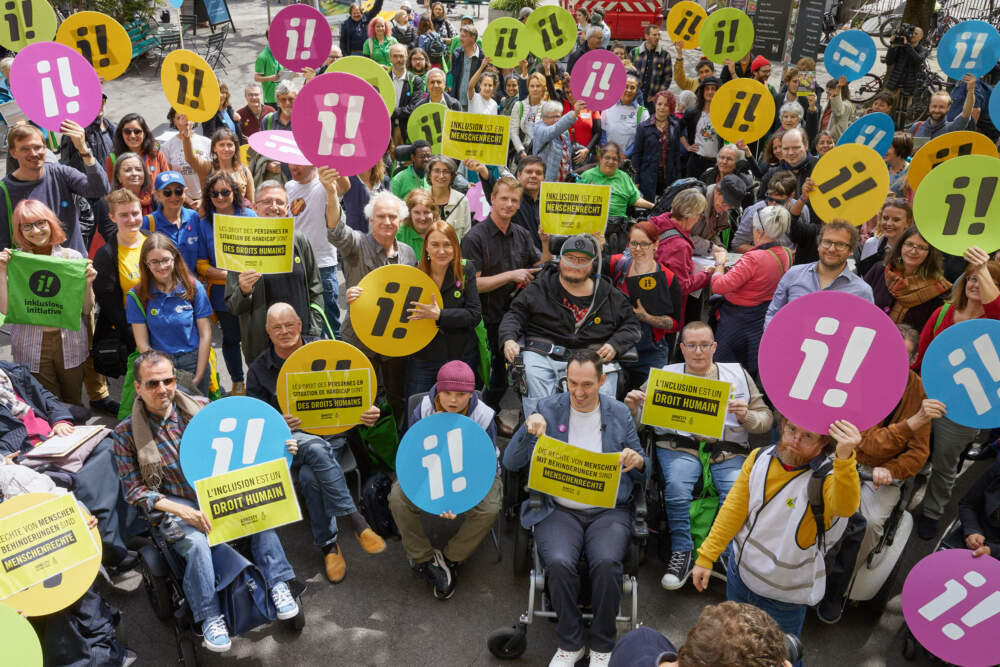 Eine Gruppe von Menschen mit und ohne Beeintr&auml;chtigung. Sie halten Schilder hoch mit einem kleinen i und einem Ausrufezeichen. Dazu Schilder, auf denen steht: Inklusion ist ein Menschenrecht.