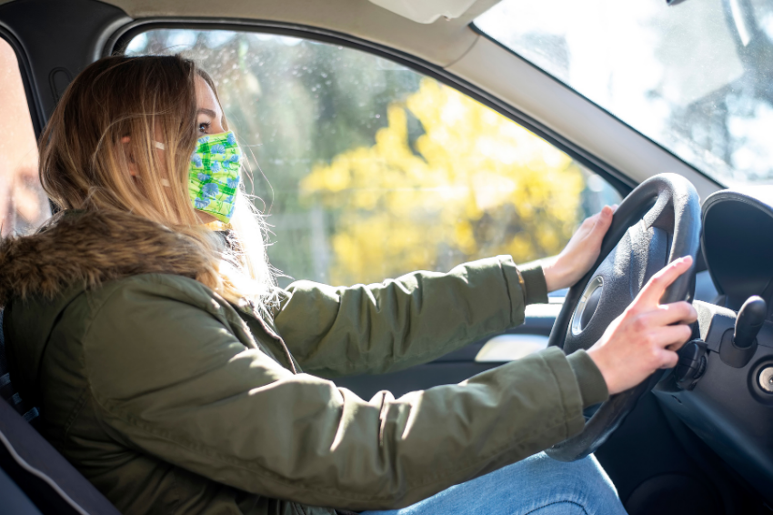 Eine Frau sitzt am Steuer eines Autos. Sie trägt eine Schutzmaske.