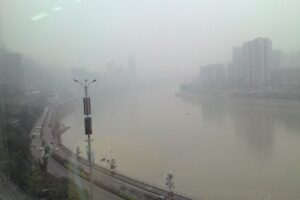Ein breiter Fluss fliesst durch eine Stadt. Smog verh&uuml;llt die Geb&auml;ude am Ufer.