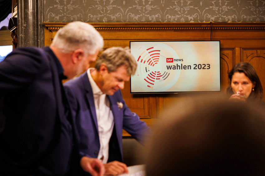 Ein Bildschirm zeigt die Sitzverteilung im Nationalrat. Daneben steht: SRF news Wahlen 2023. Vor dem Bild stehen zwei M&auml;nner und schauen sich etwas an, das vor ihnen liegt.