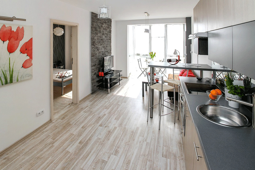 Wir blicken in eine K&uuml;che. Rechts ist die K&uuml;chenzeile mit Abwaschbecken und Herd. Dahinter kommt ein hoher Tisch mit Barhockern. Und noch weiter hinten eine Sofa und an der Wand gegen&uuml;ber ein Fernseher. Rechts schauen wir durch eine T&uuml;r ins Schlafzimmer.
