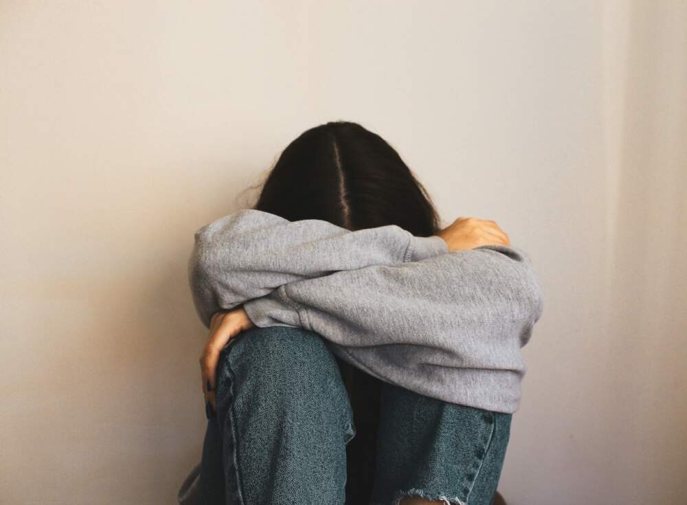 Eine Frau sitzt mit angezogenen Knien am Boden. Sie hat die Arme &uuml;ber den Knien verschr&auml;nkt. Ihr Kopf hat sie zwischen die verschr&auml;nkten Arme gelegt. Man sieht nur ihren Hinterkopf.