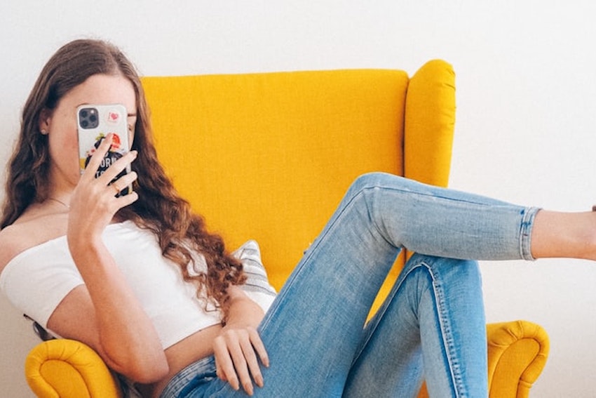 Eine junge Frau mit braunen langen Haaren trägt eine Jeans und ein weisses T-Shirt. Sie sitzt in einem gelben Sessel. Ihr Gesicht sieht man nicht. Sie hält ihr Handy vor ihr Gesicht und fotografiert sich selbst.